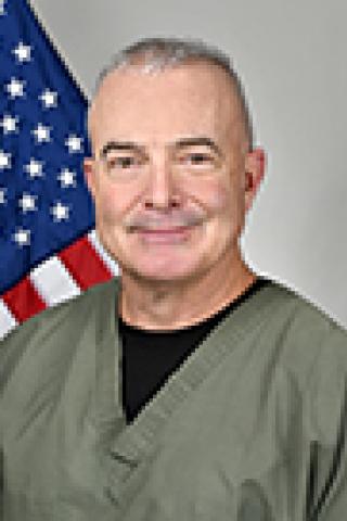 Man in green scrubs and black shirt underneath, smiling with American flag behind him.