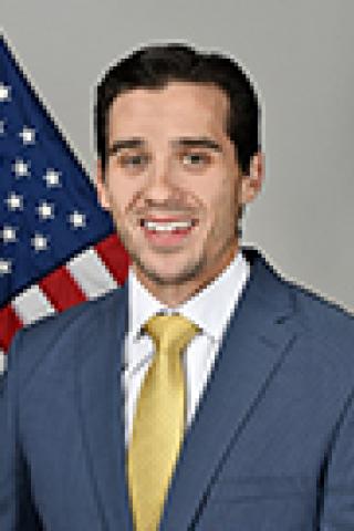 Man in blue suit and yellow tie smiles in front of American flag.