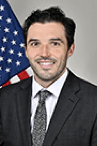 Man in a suit and tie smiling with an American flag behind him.