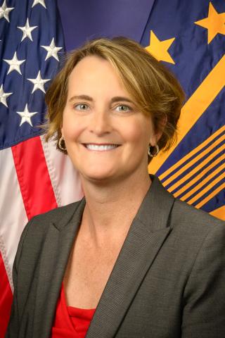 Woman in professional attire smiles in front of American and agency flags.