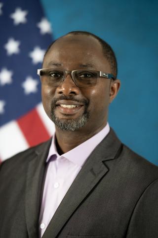 Smiling man in suit and purple shirt stands in front of US flag.
