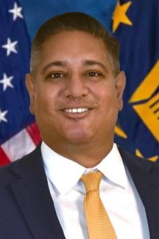 Man in a business suit and yellow tie smiles in front of flags.