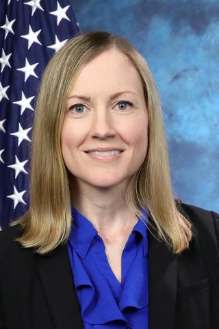 Woman with blonde hair wearing black blazer and blue shirt, smiling in front of US flag.