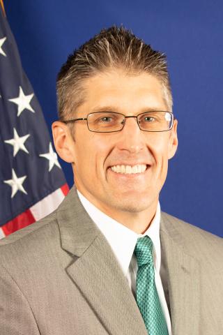 Man in suit and green tie smiling with US flag in background.