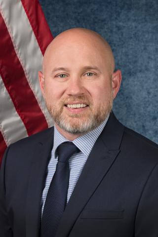 A man in a suit smiles in front of an American flag.