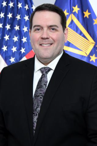 Derek Coughenour in a black suit and tie standing in front of the U.S. and SES flags.