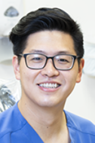 Man with glasses, short black hair, and blue scrub top smiling.