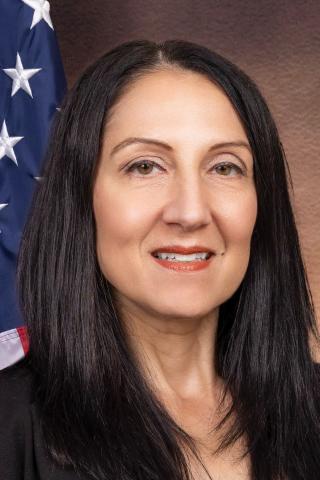 A smiling woman with long black hair stands in front of an American flag.