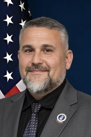 Man in gray suit and tie smiling in front of American flag.