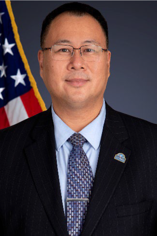 A man in a suit and tie stands in front of an American flag.
