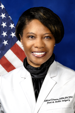A smiling African American woman in a white lab coat with a black scarf, standing in front of an American flag.