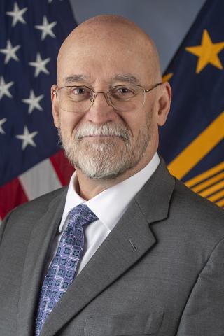 Man in a gray suit, blue tie, and glasses stands in front of the US and NSA flags.