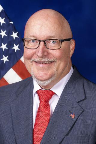 Bald man in a gray suit and red tie smiling in front of an American flag.