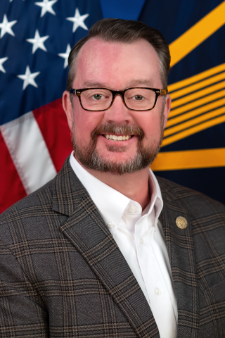 Man in suit and white shirt smiling, with American flag and blue and yellow flag in background.