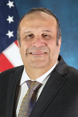A smiling man in a suit and tie stands in front of the American flag.
