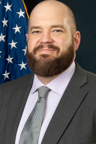 A bald man with a beard in a suit and tie, standing in front of an American flag.