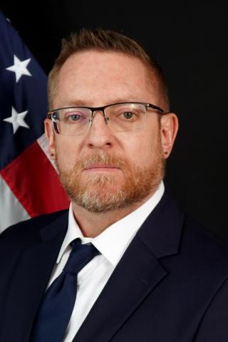 Man in formal suit and tie with American flag behind him, staring at the camera.