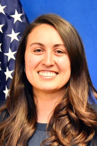 Woman with long brown hair smiling in front of an American flag.