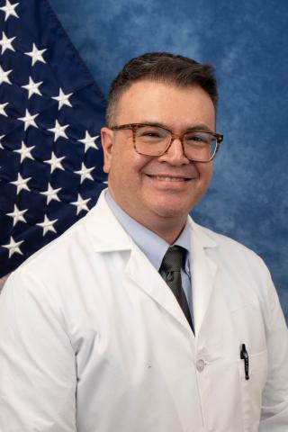 A smiling man in white lab coat, tie, and glasses stands in front of an American flag.