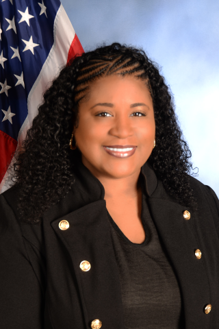 Woman with braided hair, smiling, in black attire, with an American flag behind her.