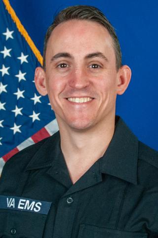 Man in black VA EMS uniform smiles in front of American flag.