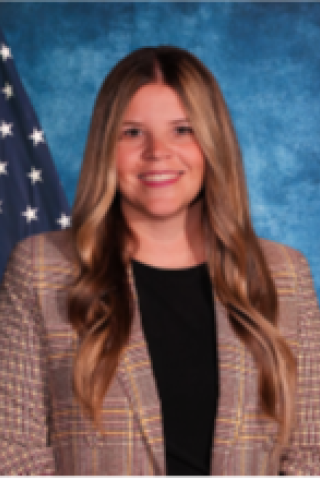 Woman with long blonde hair, wearing brown jacket and black shirt, standing in front of an American flag.