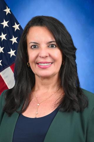 Woman with black hair and green blazer smiling in front of an American flag.