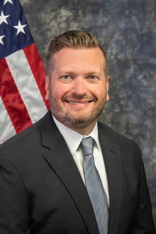 Man in a black suit and blue tie smiling next to an American flag.