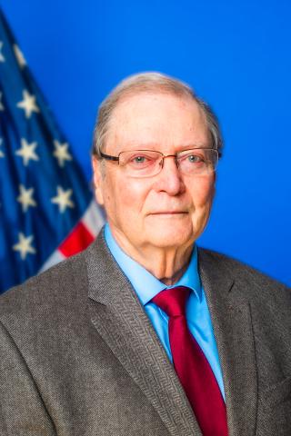 Older man in suit and red tie stands in front of American flag.