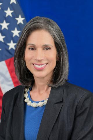 Woman with gray hair wearing blue shirt and black blazer smiling in front of American flag.
