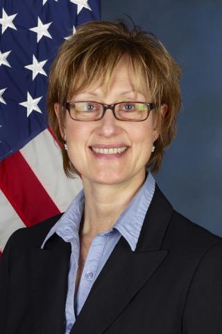 Woman in glasses and professional attire smiling in front of an American flag.