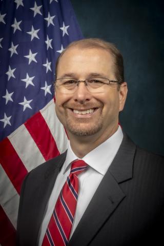 Man in suit and red striped tie smiling in front of US flag.