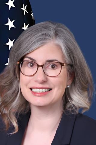 Smiling woman with gray hair wearing glasses and black blazer in front of USA flag.