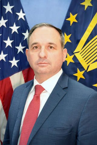 Man in suit and red tie standing in front of American and NATO flags.