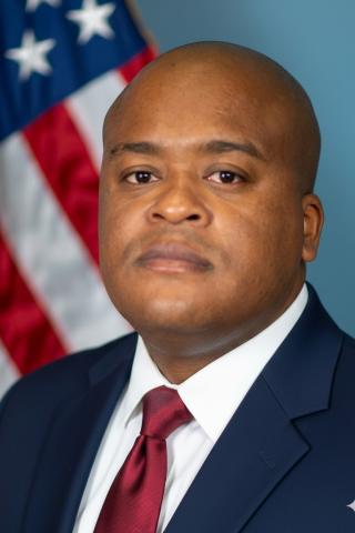 Man in a navy suit and red tie stands in front of an American flag.