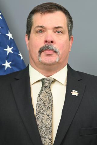 Man in formal attire with mustache, American flag in background.