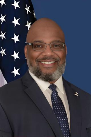 Bald man in a suit and tie smiles in front of an American flag.