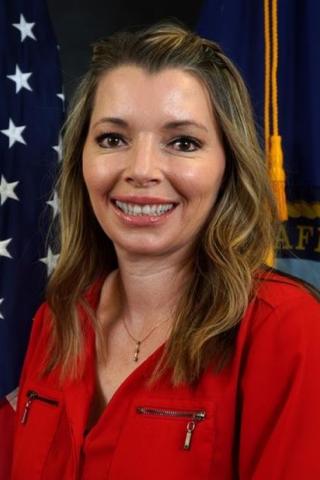 Woman in red shirt smiling in front of American and Department of State flags.