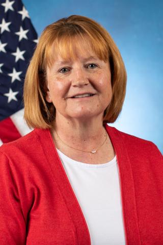 Portrait of smiling woman wearing red cardigan, white shirt, and necklace, with USA flag in background.