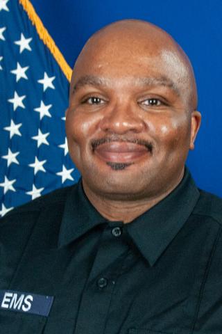 Man in black uniform with "VA EMS" patch smiling in front of a US flag.