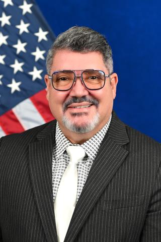 Man in a black suit and white tie, smiling with an American flag in the background.
