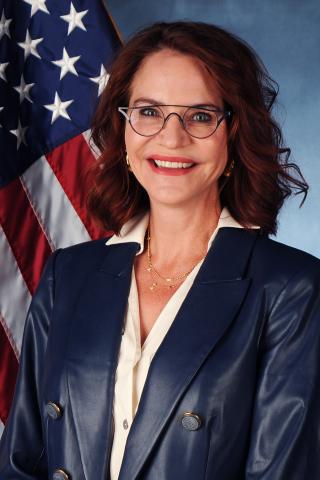 Portrait of a woman in a blue suit, smiling in front of an American flag.