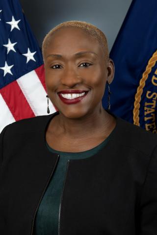 Woman with short hair in black jacket and green shirt smiles against US flag and flag with white stars on blue background.