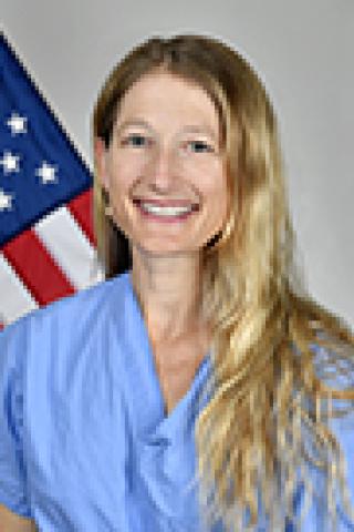 Smiling blonde woman in blue scrubs in front of an American flag.