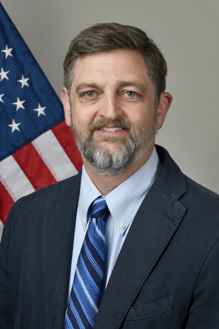 Man with a beard wearing suit and tie, smiling by an American flag.
