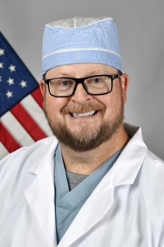 Smiling man in white lab coat, blue scrub top, and surgical cap with American flag behind him.