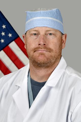 Male doctor in white lab coat and blue cap standing in front of the American flag.