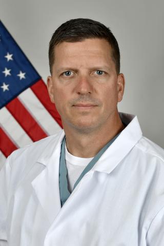 Man in white lab coat and green stethoscope stands before an American flag.