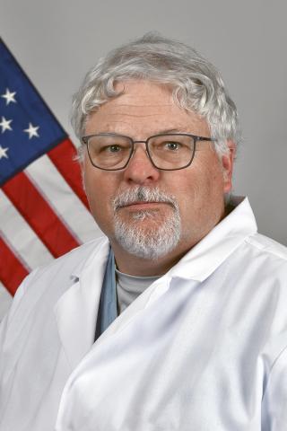 Man in white lab coat with grey hair and glasses standing before American flag.
