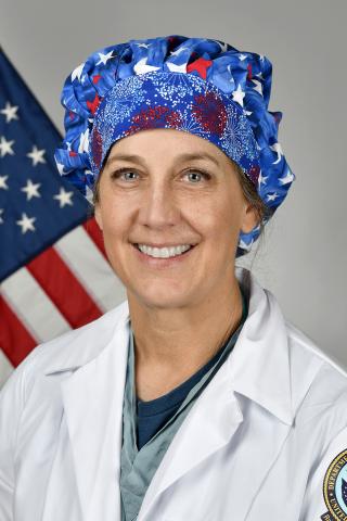 Female doctor in white lab coat and patriotic scrub cap standing in front of an American flag.